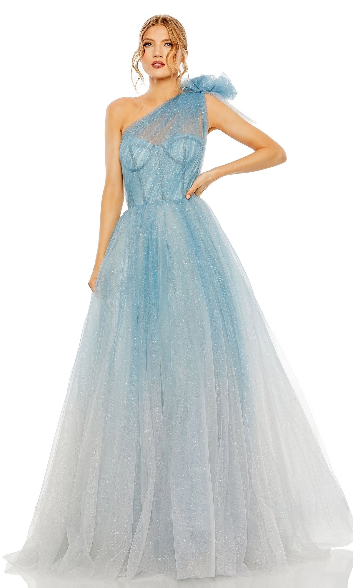 A woman stands against a white background wearing the Long Formal Dress 20552 by Mac Duggal— a light blue, one-shoulder ball gown featuring a floor-length tulle overlay and a large shoulder bow.